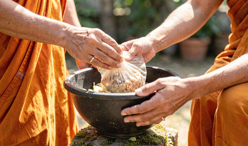 Hands placing a wrapped food offering into a monk's alms bowl on a bright morning outdoors.
